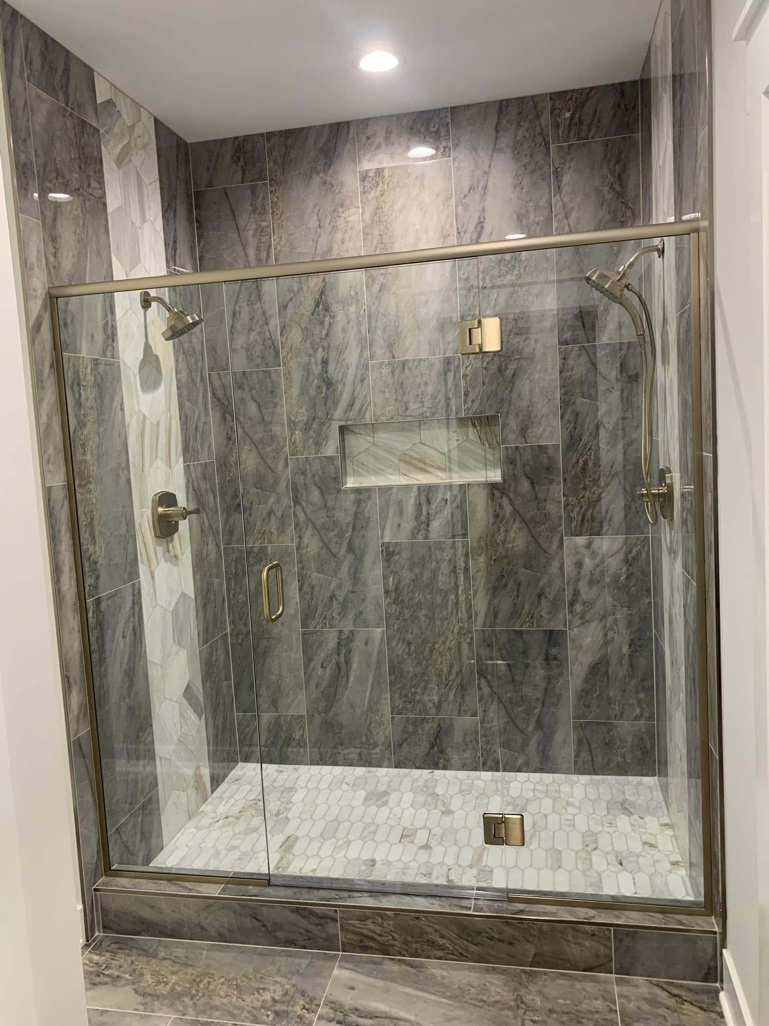 A frameless glass shower enclosure with two shower heads and large marble-look wall tile.