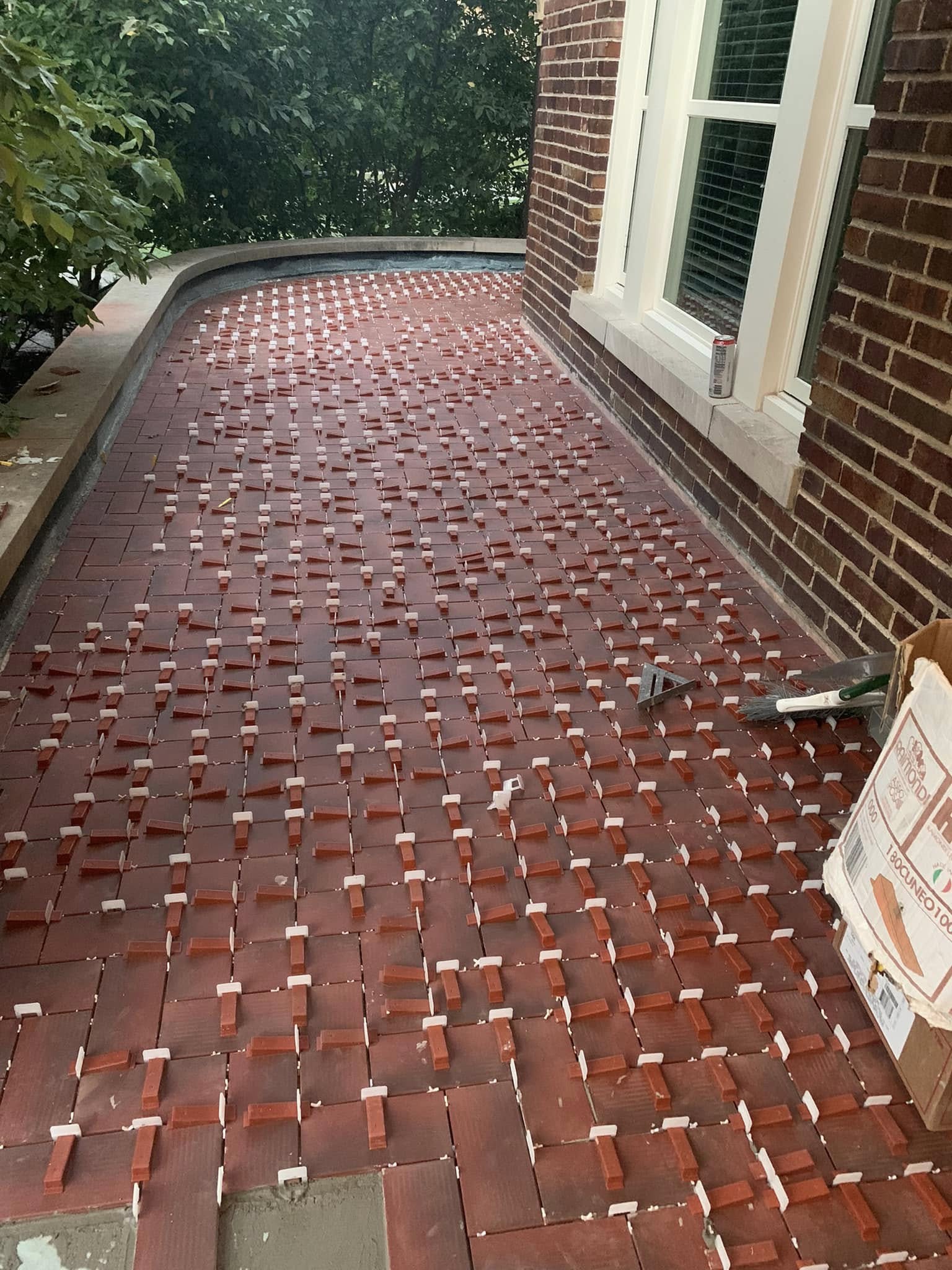 A patio part-way through installation: red brick pavers set in a pattern with white spacers between them, a trowel and broom resting on the finished section.